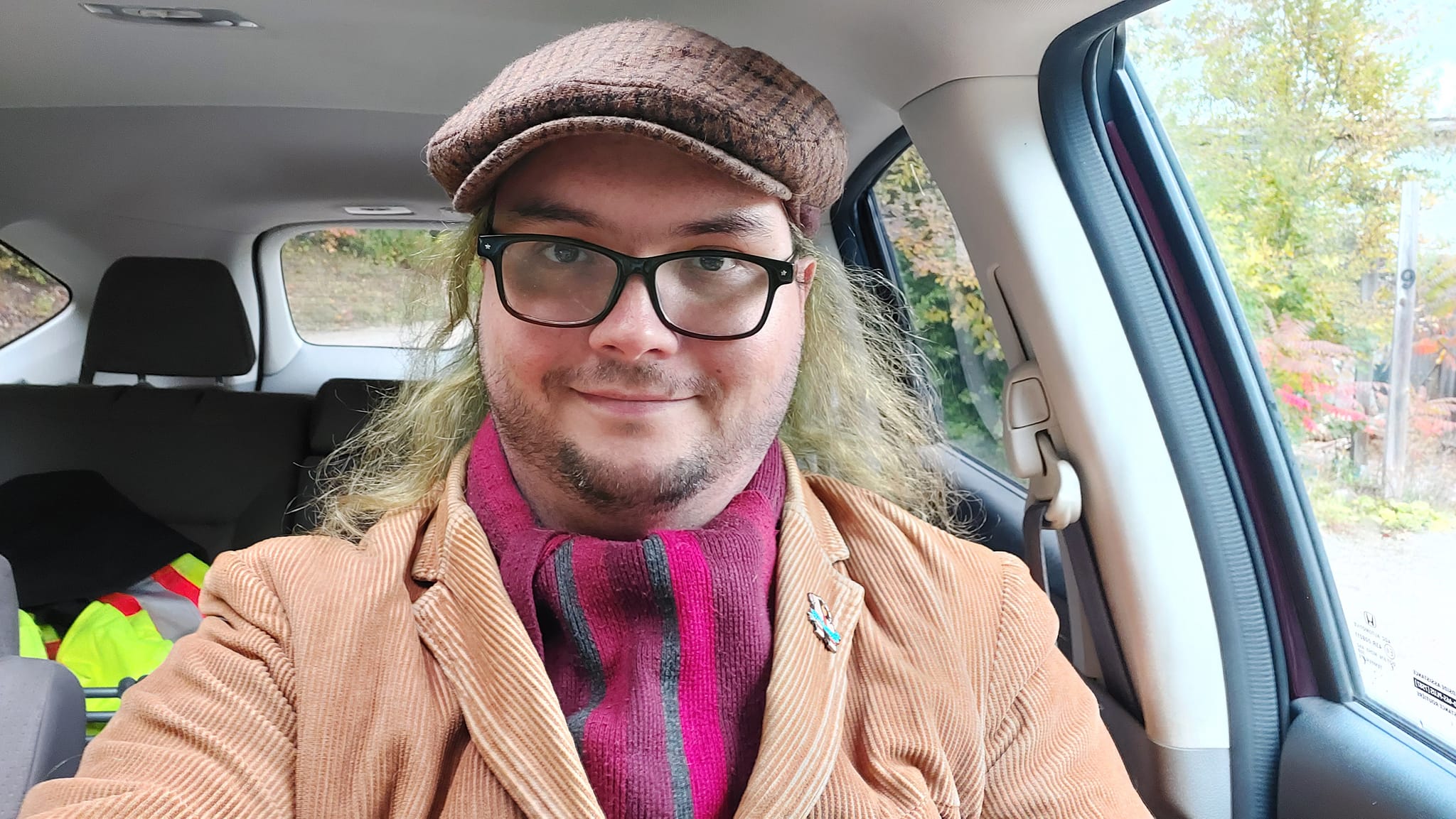 A man with long green hair wears a brown corderoy jacket and a striped red scarf, he is sitting in a car